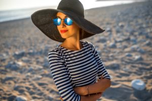 Frau sitzt mit Sonnenhut und Sonnenbrille am Strand