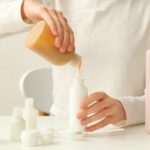 Eine Person in weißer Kleidung füllt ein Kosmetikprodukt aus einem großen Behälter in einen kleinen um. 