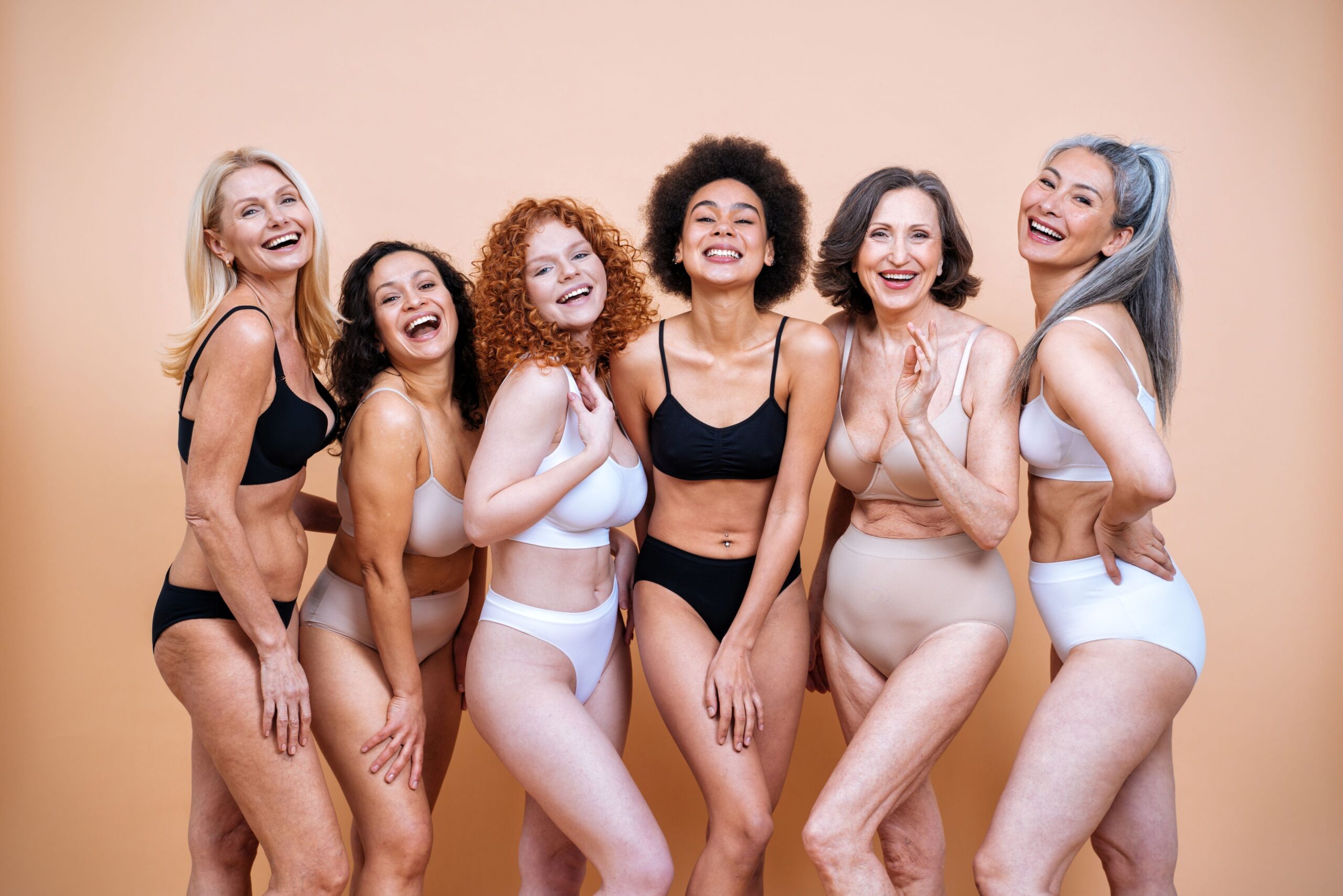 Eine Gruppe von Frauen in Unterwäsche steht vor einem pfirsichfarbenen Hintergrund. Die Frauen lächeln in die Kamera.