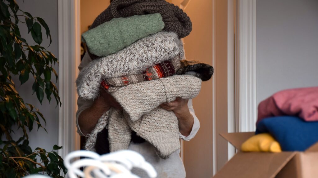 Person trägt gestapelte Pullover durch einen Raum.