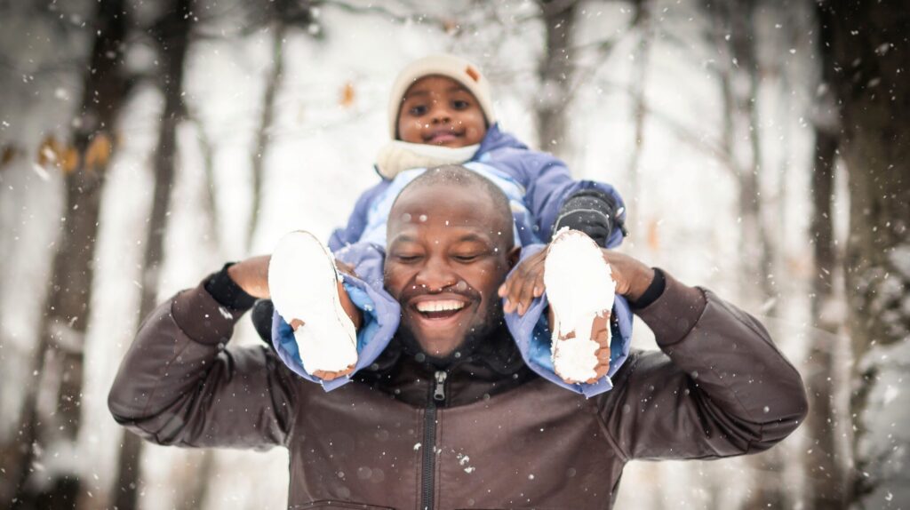 Mann in Winterjacke trägt sein Kind lachend auf den Schultern vor einem verschneiten Waldhintergrund.