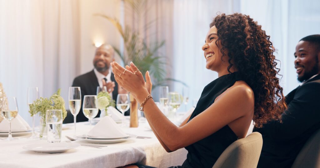 Mehrere Personen sitzen an einem festlich gedeckten Tisch. Im Vordergrund sitzt eine lächelnde Frau, die in die Hände klatscht.