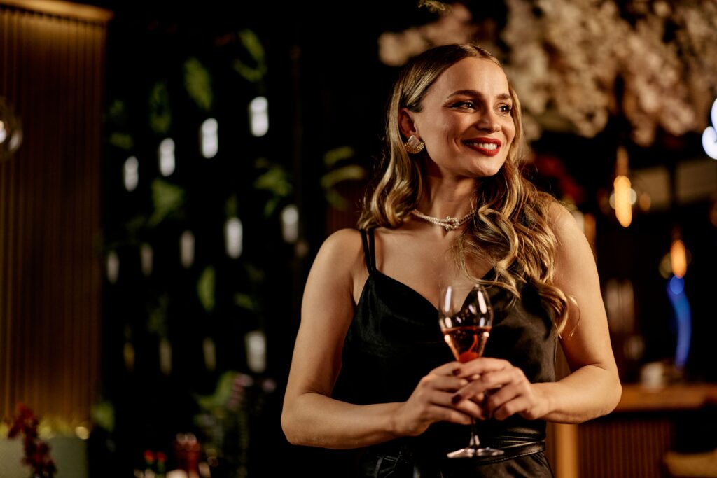 Eine junge Frau mit schwarzem Kleid und blonden Wellen hält ein Glas Sekt in der Hand und lächelt.
