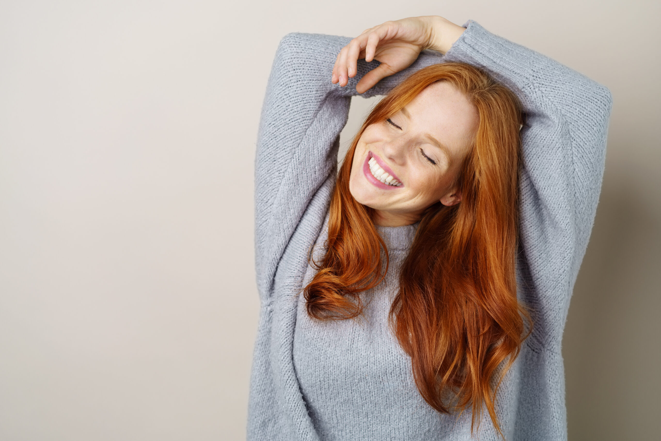 Eine rothaarige Frau mit einem grauen Pullover steht vor einem beigefarbenen Hintergrund.