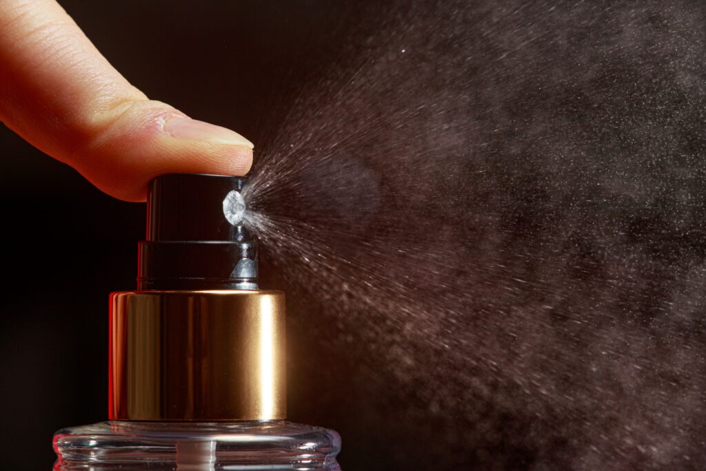 Eine Person betätigt mit dem Finger den Zerstäuber eines Parfums, wodurch das Parfum nach außen sprüht.