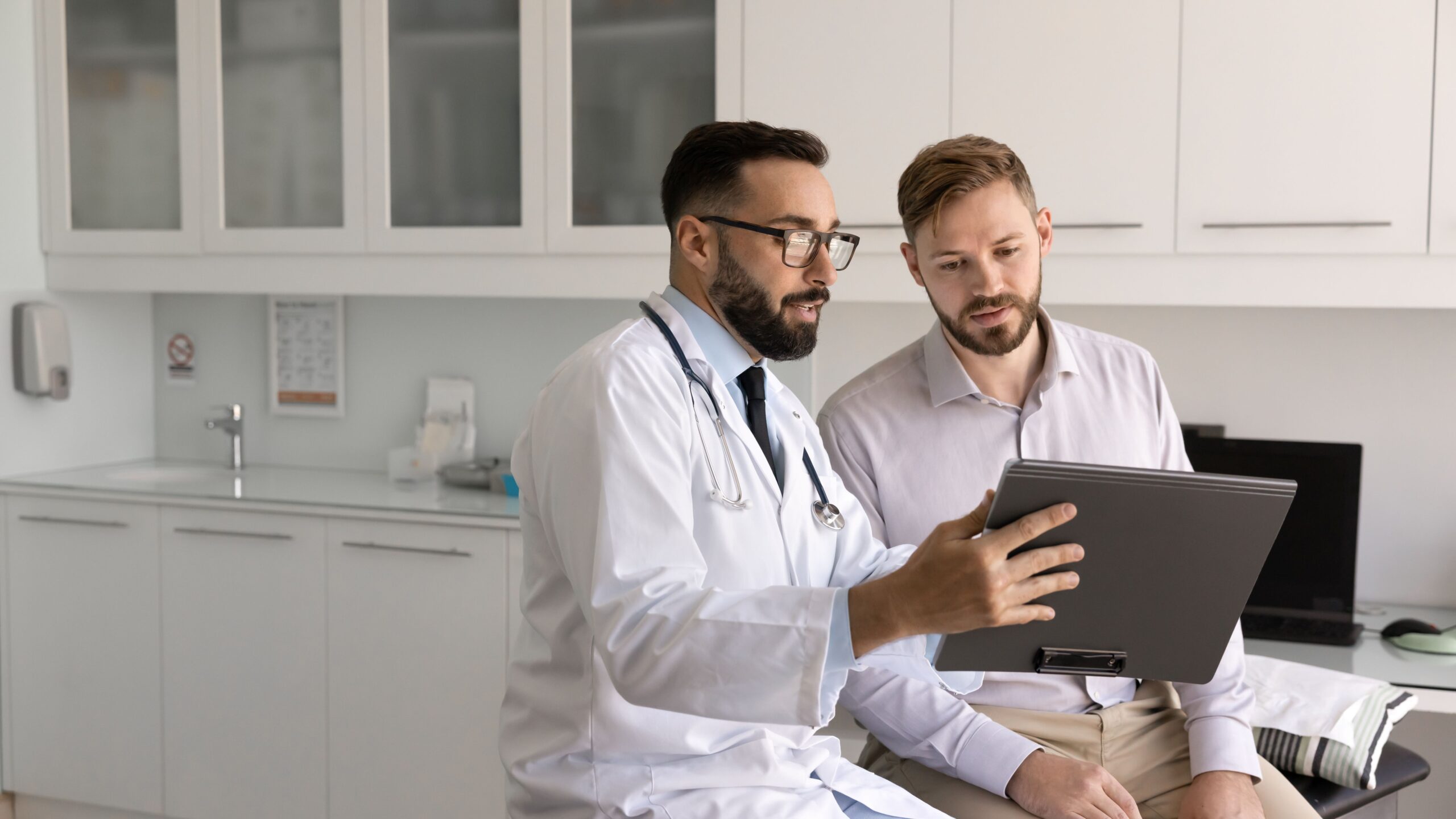 Ein Mann sitzt im Behnadlungszimmer eines Arztes. Beide blicken gemeinsam auf ein Tablet