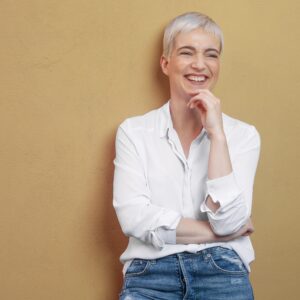Kleine Fläche, große Wirkung: So setzen Sie Accessoires bei kurzen Haaren gezielt ein Eine junge Frau mit kurzen, blonden Haaren steht lachend vor einem braunen Hintergrund.