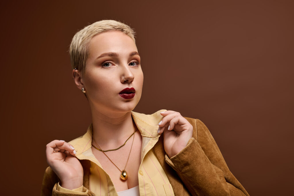 Junge Frau mit kurzen Haaren sowie auffälligem Make-up und Schmuck posiert vor einem braunen Hintergrund.