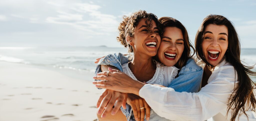 Drei Freundinnen umarmen sich und lachen. Im Hintergrund sind ein Strand und das Meer zu sehen.