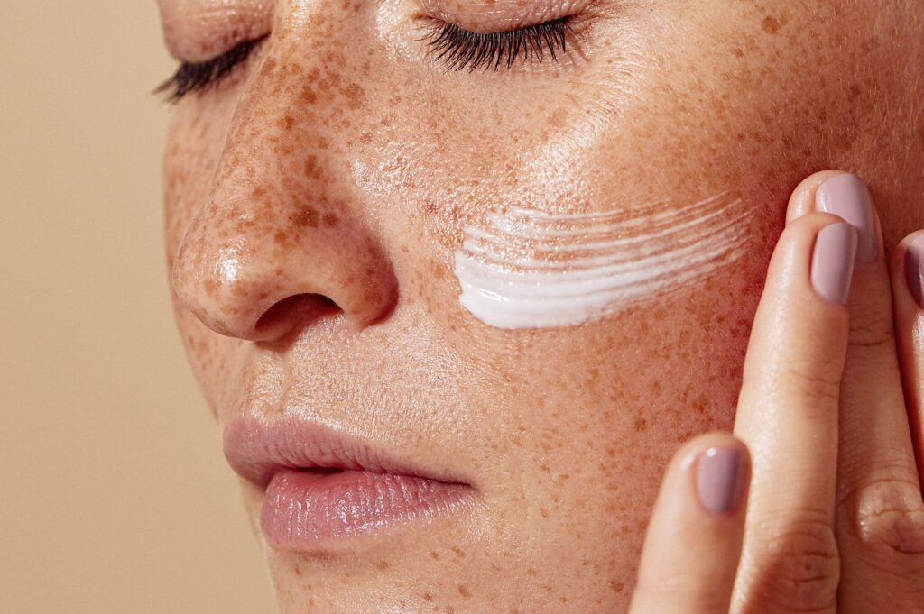 Eine junge Frau mit Sommersprossen verteilte Creme auf ihrer Wange. Ihre Augen sind dabei geschlossen.