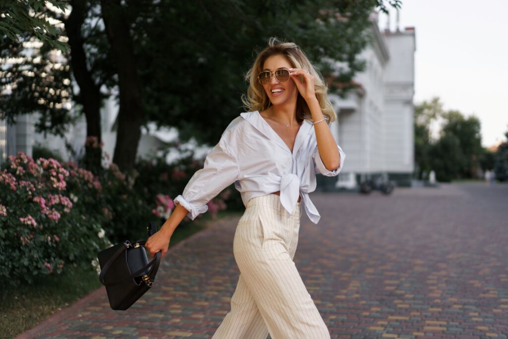 Eine Frau mit lässig, elegantem Outfit, Sonnenbrille und strahlendem Lächeln geht über eine Straße.