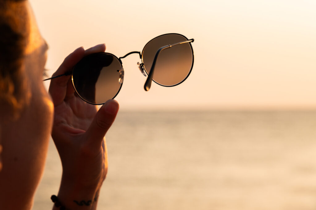 Person hält eine Sonnenbrille vor sich, im Hintergrund sind Meer und Abendlicht zu sehen.