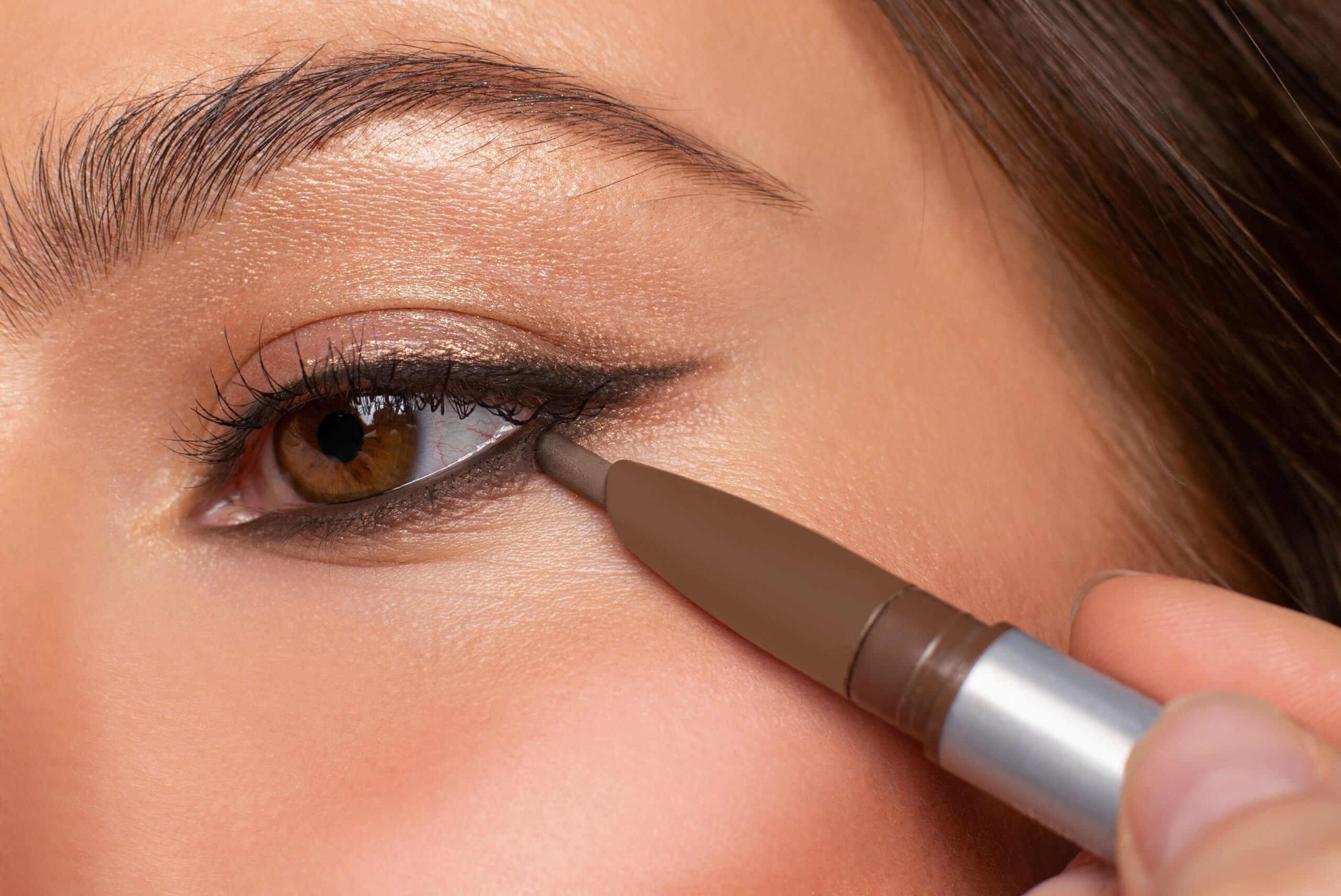 Ein Auge mit bräunlich schimmerndem Augen-Make-up ist in Nahaufnahme zu sehen, während der untere Lidstrich nachgezogen wird.