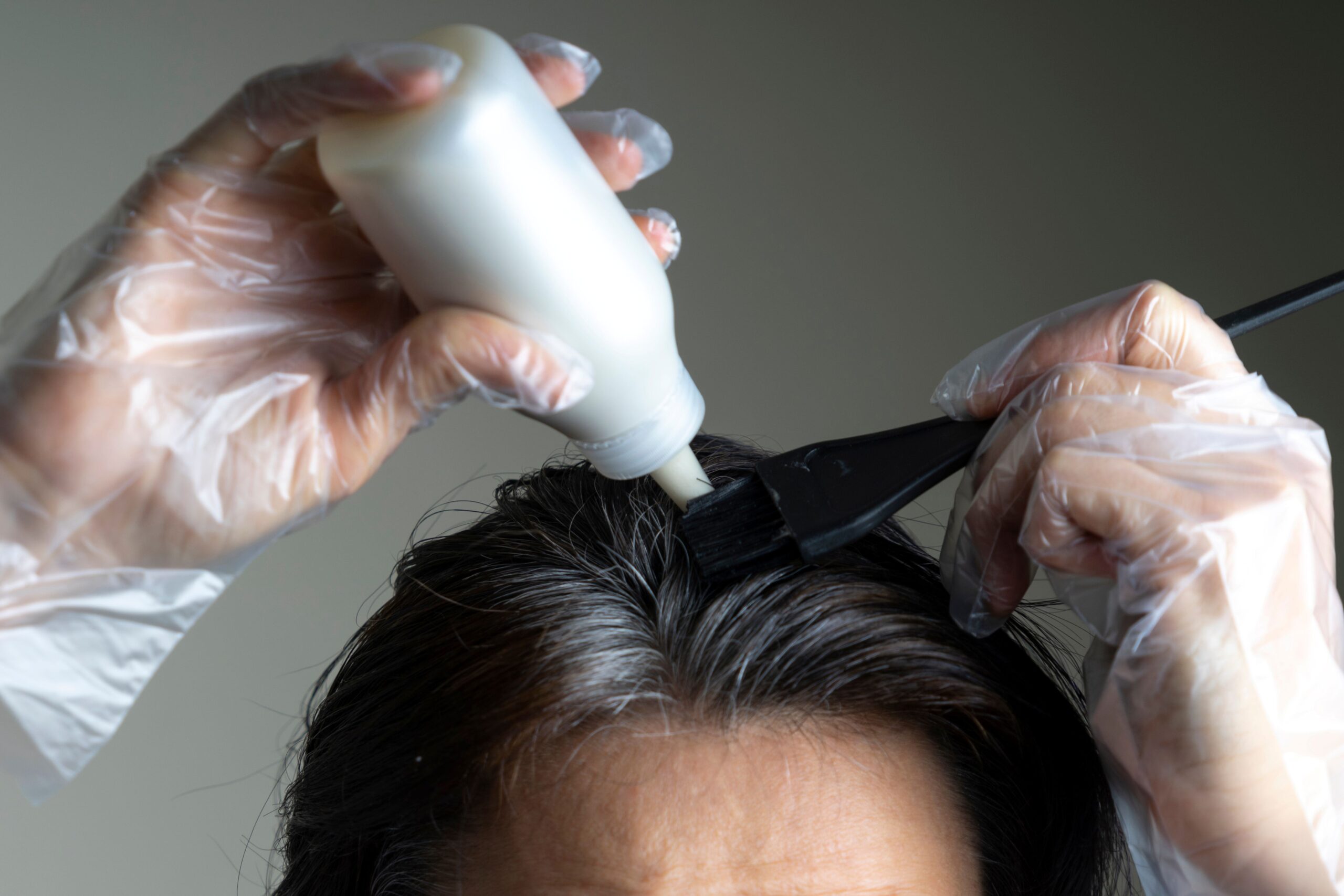 Eine Frau trägt Haarfarbe auf den grauen Ansatz ihrer Haare auf und trägt dabei Plastikhandschuhe.